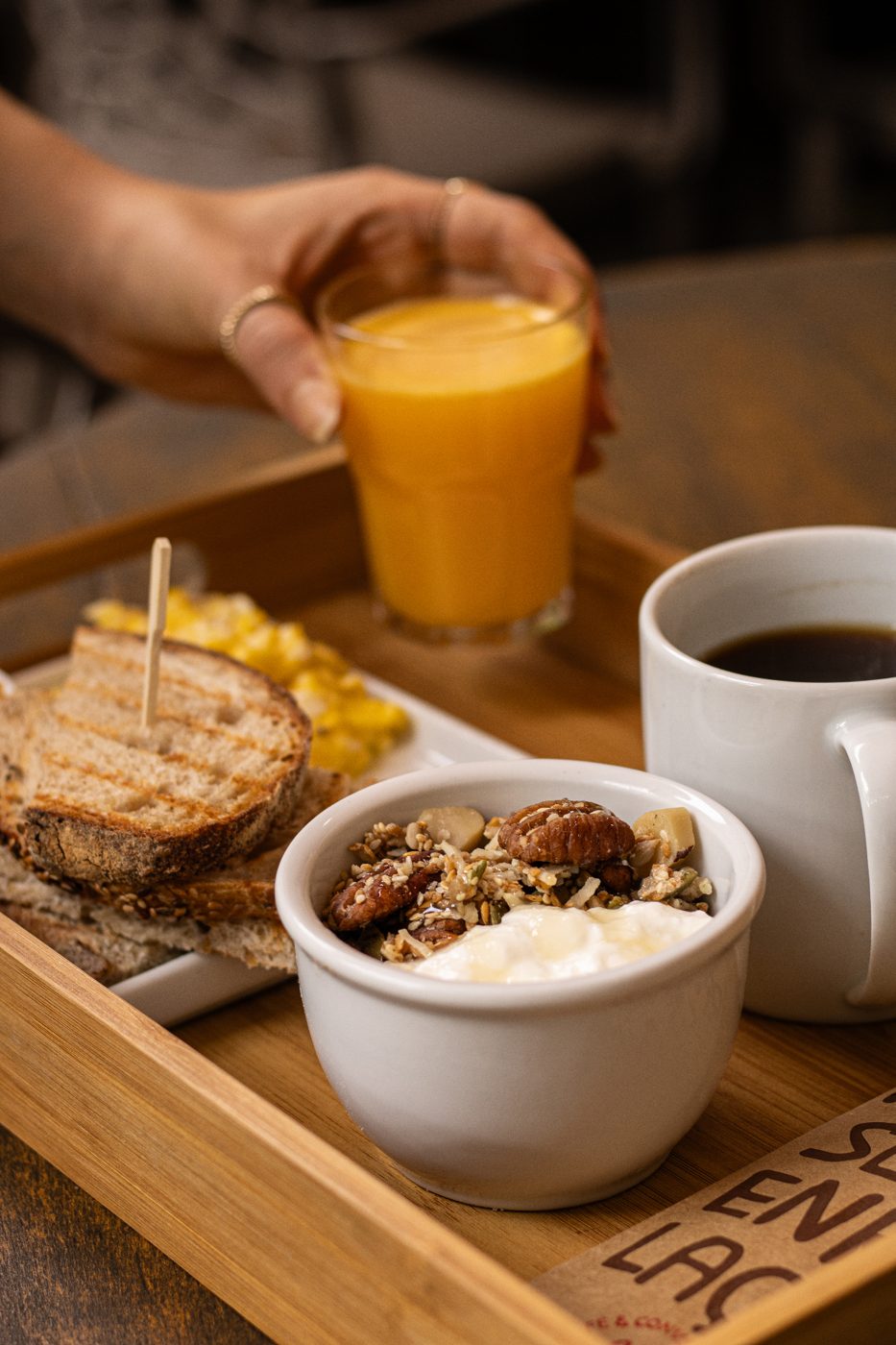 Bandeja de madeira do Yarn com torradas rústicas, ovos mexidos, tigelinha de iogurte com granola e nozes, xícara de café preto e copo de suco de laranja sendo servido.