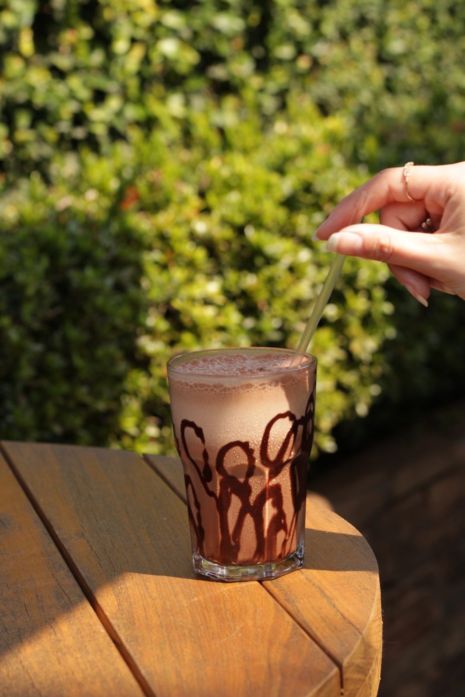 Mão mexendo com canudo uma bebida gelada de chocolate em copo alto com calda desenhada nas bordas, sobre mesa de madeira e jardim verde ao fundo.