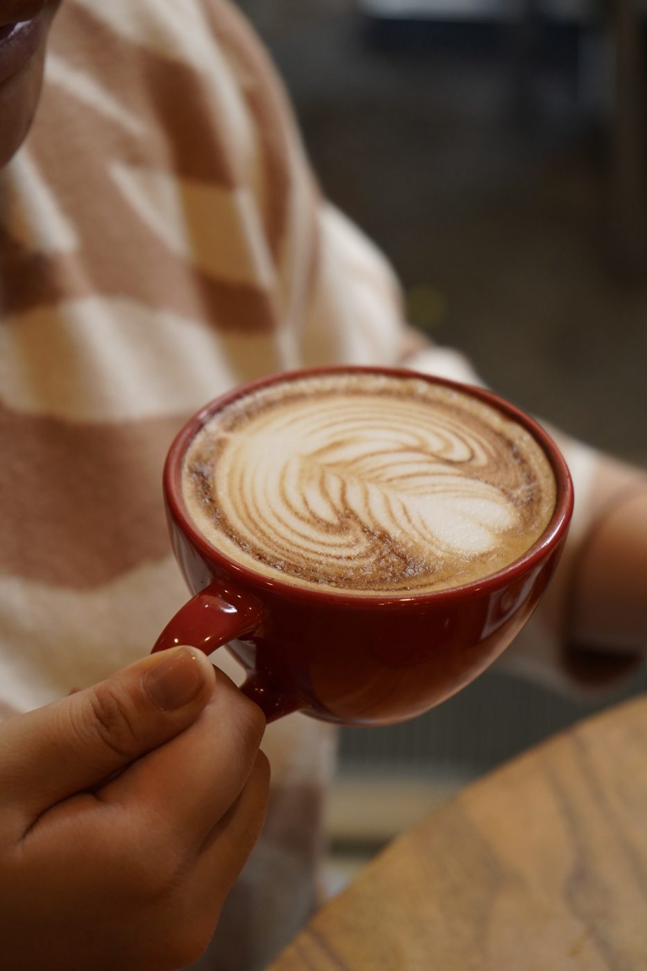 Mão segurando xícara vermelha de cappuccino com latte art em rosetta, servido no balcão do Yarn.