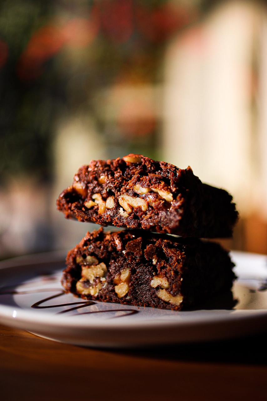 Dois pedaços de brownie de chocolate com nozes empilhados sobre prato branco riscado com fio de calda, iluminados por luz quente de fim de tarde.