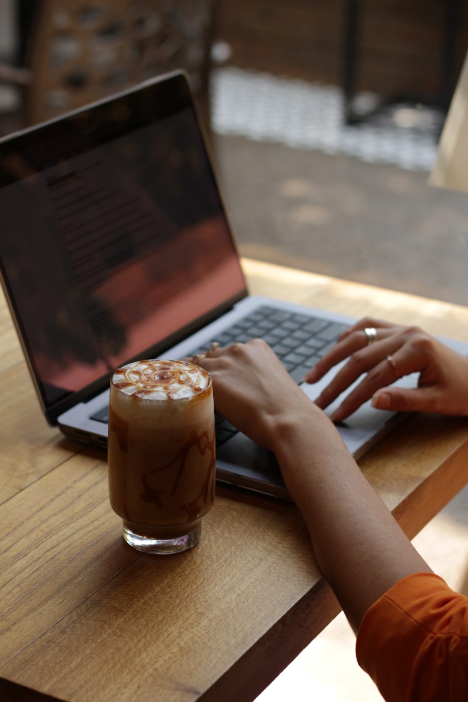 Mãos digitando em notebook sobre mesa de madeira do Yarn, ao lado de café gelado com calda de caramelo em copo alto e luz natural do salão ao fundo.